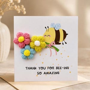 May include: A white greeting card featuring a cartoon bee illustration and three dimensional yarn flowers in pink, yellow, and blue. The card includes the printed text, THANK YOU FOR BEE-ING SO AMAZING, surrounded by small gold star accents.