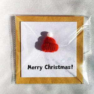 May include: A Christmas card featuring a small, knitted red hat with a white pom-pom. The card is set against a white background with the words "Merry Christmas!" in black text. The card is encased in a clear plastic sleeve.