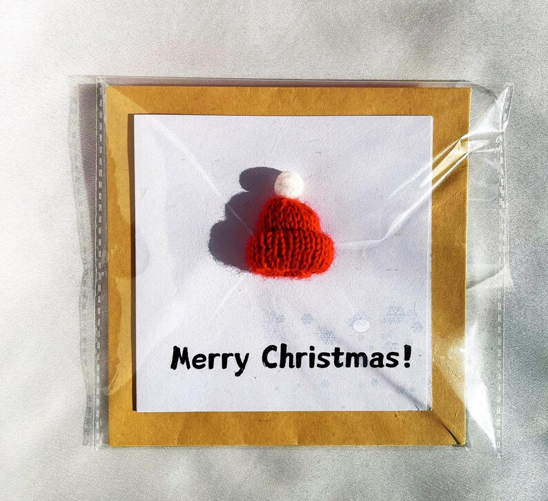 May include: A Christmas card featuring a small, knitted red hat with a white pom-pom. The card is set against a white background with the words "Merry Christmas!" in black text. The card is encased in a clear plastic sleeve.