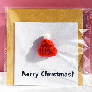 May include: A Christmas card featuring a small, red knitted Santa hat with a white pom-pom. The card has the text "Merry Christmas!" in black, with a gold backing and is encased in a clear plastic sleeve. The background is a solid pink.
