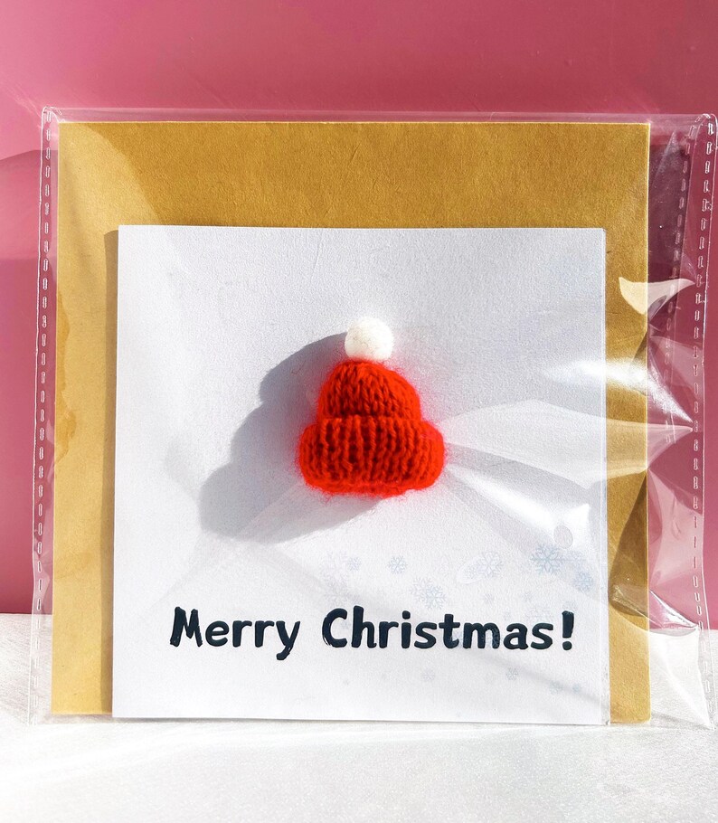 May include: A Christmas card featuring a small, red knitted Santa hat with a white pom-pom. The card has the text "Merry Christmas!" in black, with a gold backing and is encased in a clear plastic sleeve. The background is a solid pink.