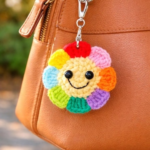 Peut inclure: Un porte-clés en forme de fleur crochetée aux couleurs vives avec un visage souriant. La fleur a des pétales rouges, roses, oranges, jaunes, verts, bleus et violets. Le porte-clés est attaché à un sac à main en cuir marron avec un fermoir argenté.