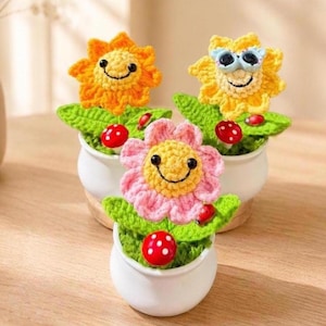 May include: Three crocheted flower pot decorations. Each pot features a smiling flower with a yellow centre and petals in orange, yellow, and pink. Green leaves and red ladybirds are also included. The pots are white.