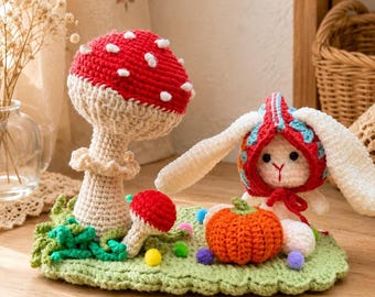 May include: A crochet display featuring a large red and white mushroom, a smaller red mushroom, a white rabbit wearing a red and blue patterned hood, and an orange pumpkin, all set on a light green crocheted base with small colorful pom-poms.