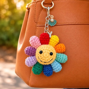 May include: A vibrant crochet sunflower keyring with a smiling face, attached to a tan leather handbag. The keyring features a silver clasp, a small charm, and a letter "Y" charm. The sunflower has petals in various colours.