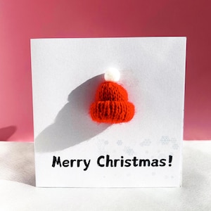 May include: A white greeting card with a 3D knitted red Santa hat and the text "Merry Christmas!" in black. The hat has a white pom-pom on top. The background is a gradient of pink and white.