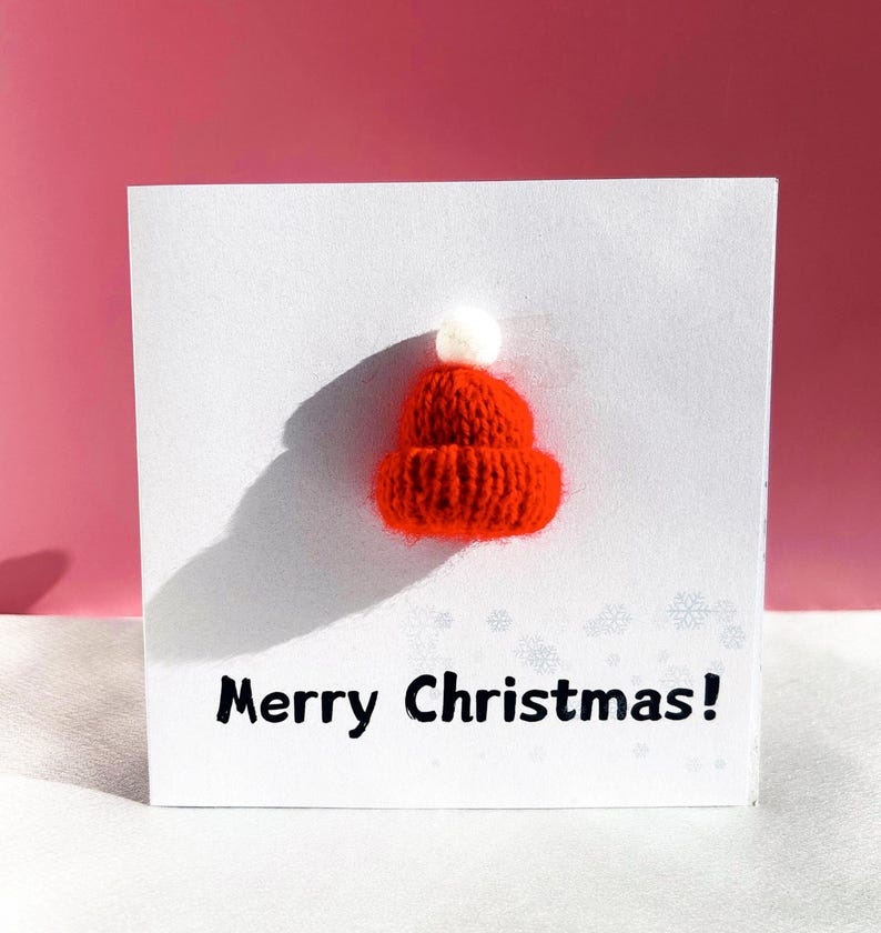 May include: A white greeting card with a 3D knitted red Santa hat and the text "Merry Christmas!" in black. The hat has a white pom-pom on top. The background is a gradient of pink and white.