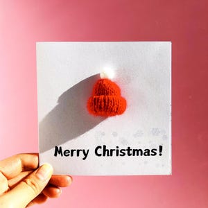 May include: A greeting card with a small, knitted red hat with a white pom-pom. The card reads "Merry Christmas!" in black text. The background is a solid pink color.
