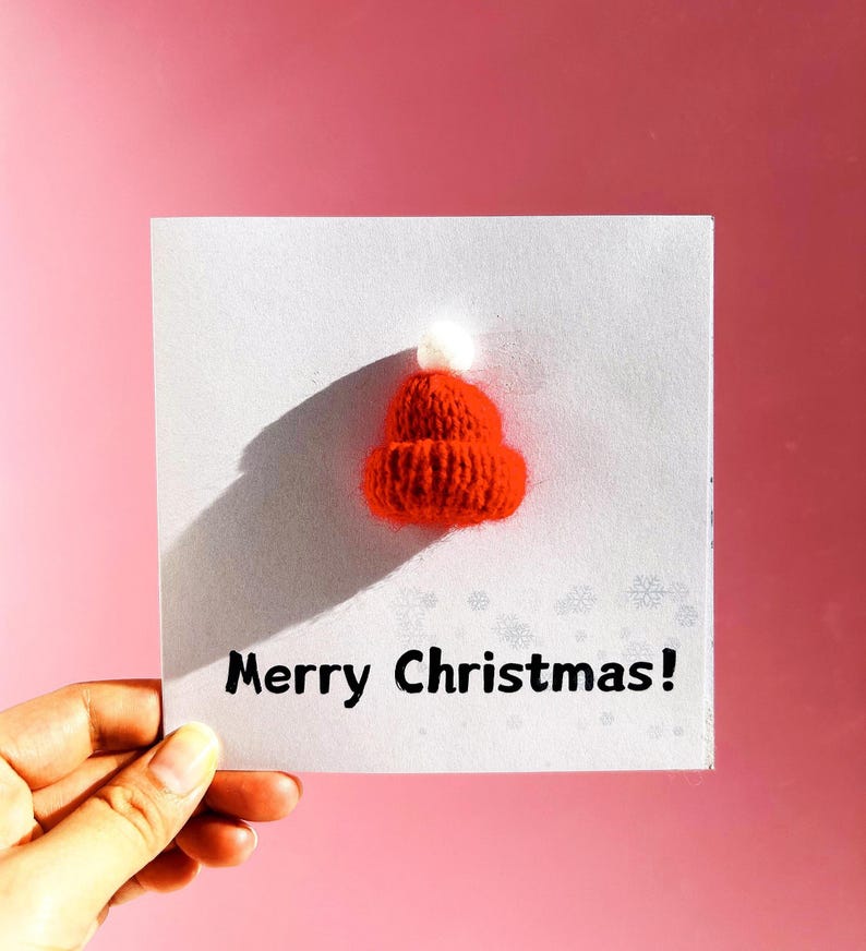 May include: A greeting card with a small, knitted red hat with a white pom-pom. The card reads "Merry Christmas!" in black text. The background is a solid pink color.