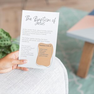May include: A person holds a white card with the title "The Baptism of Jesus" in cursive. The card includes text about the baptism and a quote from Matthew 3:13-17. A potted plant and a small table are in the background.