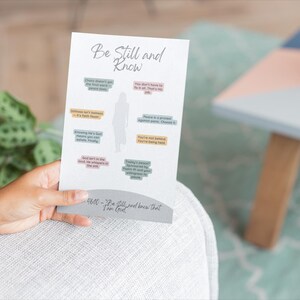 Puede incluir: Una tarjeta blanca con las palabras "Be Still and Know" y citas inspiradoras en varios colores. La tarjeta está en manos de una persona. Una planta en maceta y una mesita están en el fondo.