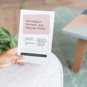 May include: A white card with the text "No weapon formed... but they be TRYIN'" in a pink box. The card also lists Bible verses. The card is held by a person.