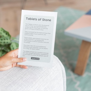 May include: A person holding a white card titled "Tablets of Stone" with text about the Ten Commandments. The card is held in front of a blurred background with a plant and a small table.