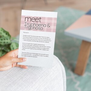 以下が含まれることがあります： ピンクのチェック柄のバナーに「meet Tryphaena & Tryphosa」というテキストが入った白いカード。カードは人が持っています。鉢植えの植物と小さなテーブルが背景にあります。