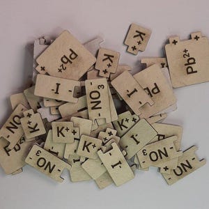 Peut inclure: Un ensemble de petites pièces de puzzle en bois de couleur claire. Chaque pièce présente des symboles et des formules chimiques, notamment Pb2+, K+, I-, et NO3-. Les pièces sont éparpillées, suggérant un thème scientifique ou éducatif.