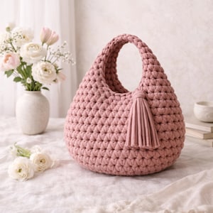 May include: A dusty rose crochet handbag with a rounded shape and a single handle. The bag features a textured stitch pattern and a matching tassel. The bag is sitting on a white surface.