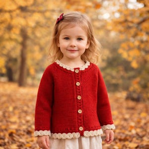 Puede incluir: Una niña pequeña con un cárdigan rojo tejido con un ribete de ganchillo color crema alrededor del cuello, los puños y el dobladillo. El cárdigan tiene botones de madera en la parte delantera. El fondo es una escena otoñal borrosa.