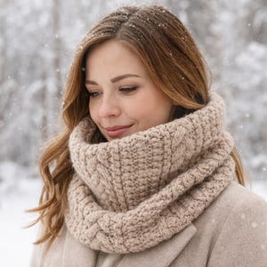 Op de afbeelding: Beige kabelgebreide col sjaal. De sjaal is gemaakt van dik, zacht garen en is ontworpen om om de nek te worden gedragen. De vrouw op de foto draagt de sjaal buiten in een besneeuwde omgeving. De sjaal heeft een neutrale kleur en past bij veel outfits.