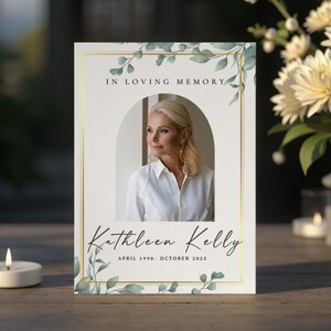 Op de afbeelding: Een herdenkingskaart met een foto van een vrouw in een wit overhemd, omlijst door een gouden rand en groen blad. De tekst luidt "In Loving Memory" en "Kathleen Kelly, april 1998 - oktober 2025". Een brandende kaars en bloemen staan op de achtergrond.