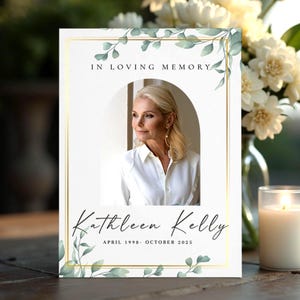 Può includere: Un cartello commemorativo con la foto di una donna in camicia bianca, incorniciata da un bordo dorato e da un fogliame verde. Il testo recita "In Loving Memory" e "Kathleen Kelly, aprile 1998- ottobre 2025". Una candela accesa e dei fiori sullo sfondo.