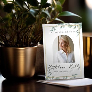 Op de afbeelding: Een herdenkingskaart met de tekst "In Loving Memory" en de naam "Kathleen Kelly". De kaart toont een foto van een vrouw en is versierd met groene bladeren en een gouden rand. Een potplant en een gouden kandelaar staan op de achtergrond.
