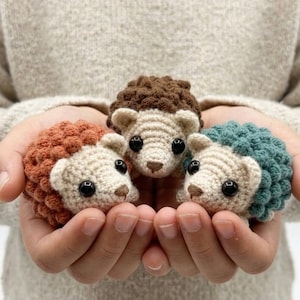 May include: Three crocheted hedgehogs in shades of brown, orange, and teal are held in cupped hands. The hedgehogs have cream-colored faces and black eyes. The spiky backs are in contrasting colors. The background is a neutral-colored sweater.