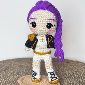 Puede incluir: Muñeca de ganchillo hecha a mano con una trenza morada, ojos negros y mejillas rosadas. La muñeca viste un atuendo blanco con detalles dorados y negros, incluyendo una chaqueta y botas. Se encuentra sobre una rodaja de madera.