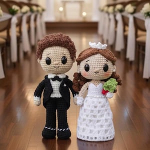 Può includere: Due bambole all'uncinetto, sposo e sposa, si tengono per mano su un pavimento di legno. Lo sposo indossa uno smoking nero con camicia bianca e papillon. La sposa indossa un abito bianco e tiene un piccolo bouquet.