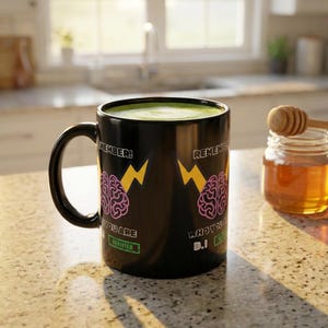 Op de afbeelding: Zwarte keramische mok met een groene vloeistof erin. De mok heeft een ontwerp met een roze hersenen, gele bliksemschichten en de woorden "Remember! Who You Are Verified B.I."