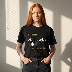 Op de afbeelding: Zwart t-shirt met een grafisch ontwerp met een pinguïn, bergen en de tekst "AI RISES STILL HERE" in groen en geel. Het ontwerp bevat een weg en een ondergaande zon.