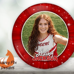 May include: Red and white round cheerleading button with a photo of a girl in a cheerleading uniform. The button has a red background with white stars and the text "Shelby JACKSON" in white.
