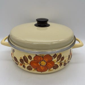 May include: Cream-colored enamel pot with lid and handles. The pot features a floral design in shades of orange and brown. The lid has a black knob. A vintage kitchenware item.