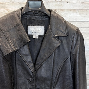 May include: Black leather jacket with a notched collar and a single-button closure. The jacket features a tailored fit and the label reads "Wilsons Leather Maxima". The jacket is hanging on a black hanger.