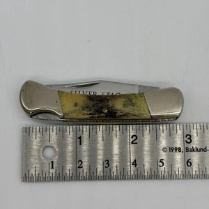 以下が含まれることがあります： クリーム色と茶色のテクスチャ加工されたハンドルと銀色の金属製ボルスターを備えた折りたたみ式ポケットナイフ。ブレードにはSILVER STAGの文字が刻印されています。ナイフは、約7.6センチメートルの長さを示す金属製の定規の上に配置されています。