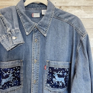 May include: A denim button-up shirt with two front pockets featuring a blue deer and snowflake design. The shirt has a classic collar, buttoned cuffs, and a Levi's tag. The shirt is a medium wash denim.