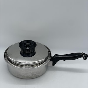 Op de afbeelding: Een roestvrijstalen steelpan met bijpassend deksel en een zwarte handgreep. Het deksel heeft een zwarte knop in het midden. De pan heeft een gepolijste metalen afwerking en een stevige zwarte handgreep met een metalen lus aan het uiteinde.
