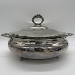 Könnte beinhalten: Eine silberbeschichtete Servierschale mit Glaseinsatz und Deckel. Die Schale hat verzierte Griffe und einen dekorativen Griff am Deckel. Die Schale steht auf drei kleinen Füßen. Das Gesamtdesign ist klassisch und elegant.