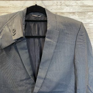 May include: A gray blazer with a notched lapel and two buttons on the cuff. The jacket is hanging on a black hanger against a light wood background. The blazer has a textured appearance.