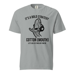 Puede incluir: Camiseta gris claro con el texto "IT'S A BOLD STRATEGY" sobre un gráfico de una cabeza de serpiente con auriculares. Debajo, el texto "COTTON (MOUTH)" y "LET'S SEE IF IT PAYS OFF FOR EM".