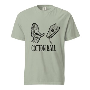 Peut inclure: T-shirt vert clair avec un motif noir et blanc de deux têtes de serpent. Le texte "COTTON BALL" est imprimé en noir en dessous. Le t-shirt est fait d'une matière douce et confortable.