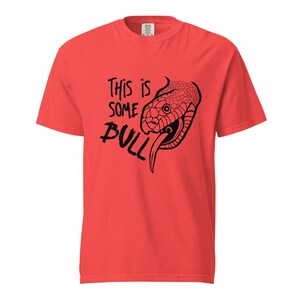 Könnte beinhalten: Rotes T-Shirt mit einer schwarzen Grafik eines Schlangenkopfes und dem Text "This is SOME BULL". Der Schlangenkopf ist detailliert mit herausgestreckter Zunge, und der Text ist in einer handschriftlichen Schriftart.