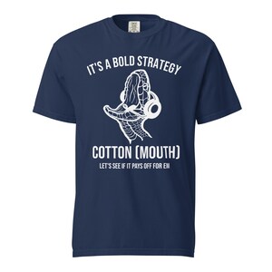 Puede incluir: Camiseta azul marino con texto blanco y un gráfico de una cabeza de serpiente con auriculares. El texto dice "IT'S A BOLD STRATEGY", "COTTON (MOUTH)" y "LET'S SEE IF IT PAYS OFF FOR EM".