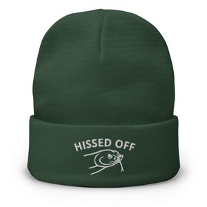 Op de afbeelding: Een donkergroene beanie met de tekst "HISSED OFF" in het wit geborduurd, vergezeld van een cartoon slang. De beanie heeft een omgeslagen manchet en is gemaakt van een zachte, gebreide stof.