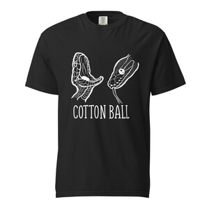 Op de afbeelding: Zwart T-shirt met een witte afbeelding van twee slangenkoppen. Eén heeft een open mond, de andere een uitgestoken tong. De tekst "COTTON BALL" is wit onder de slangenkoppen gedrukt.