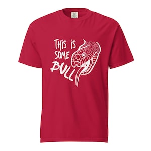 Peut inclure: T-shirt rouge avec un motif blanc représentant une tête de serpent et le texte "This is some bull". La tête de serpent est détaillée avec des écailles et une langue. Le t-shirt est de couleur rouge uni.