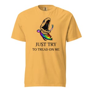 Op de afbeelding: Mosterdgeel T-shirt met een afbeelding van een slang met een regenboogtong en accenten. De tekst "JUST TRY TO TREAD ON ME" staat onder de slangillustratie. Het shirt heeft korte mouwen en een ronde hals.