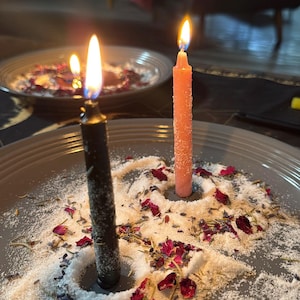 May include: Two lit candles, one black and one peach, stand on a gray plate surrounded by white granules, dried rose petals, and herbs. The candles' flames are bright against the dark background, suggesting a ritual or spiritual practice.
