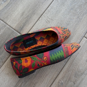 May include: A pair of Kenzo flat shoes featuring a vibrant red base with a multi-colored geometric pattern and floral motifs. The interior lining displays a matching geometric print with a black label featuring the Kenzo brand name in blue.