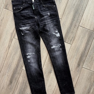May include: Black denim jeans with distressed detailing. The jeans have a button closure, pockets, and a small green tag. The denim has a faded appearance with intentional rips and wear.