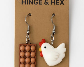 Pode incluir: Um par de brincos pendentes com uma galinha branca e uma caixa de ovos marrom com 12 ovos marrons. Os brincos estão em um cartão marrom com o texto "HINGE & HEX".